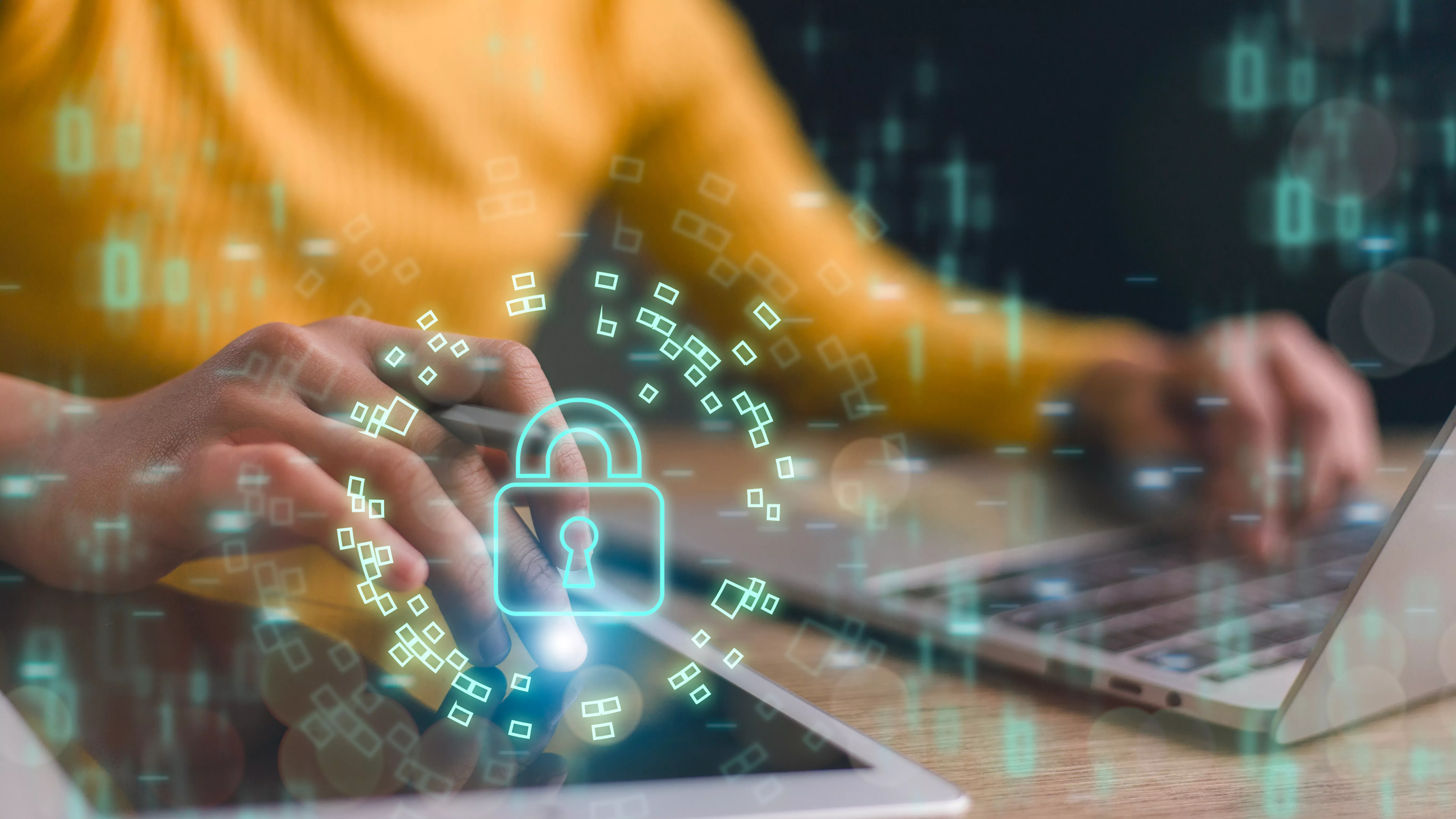 Osoba pracująca na laptopie i tablecie, na które nałożona jest świecąca, cyfrowa grafika kłódki otoczonej danymi. Symbol cyberbezpieczeństwa i ochrony danych w internecie